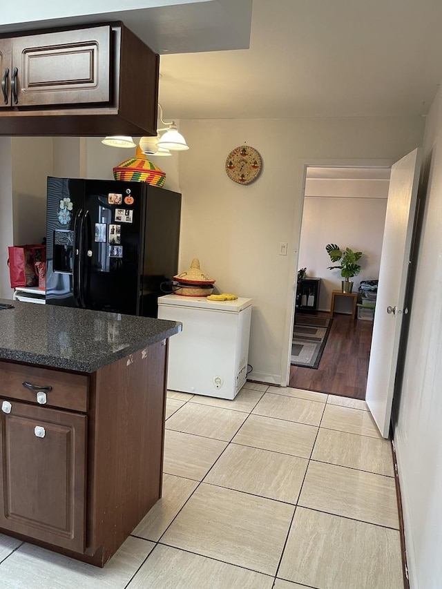 kitchen featuring refrigerator, black fridge, dark brown cabinets, light tile patterned floors, and dark stone countertops