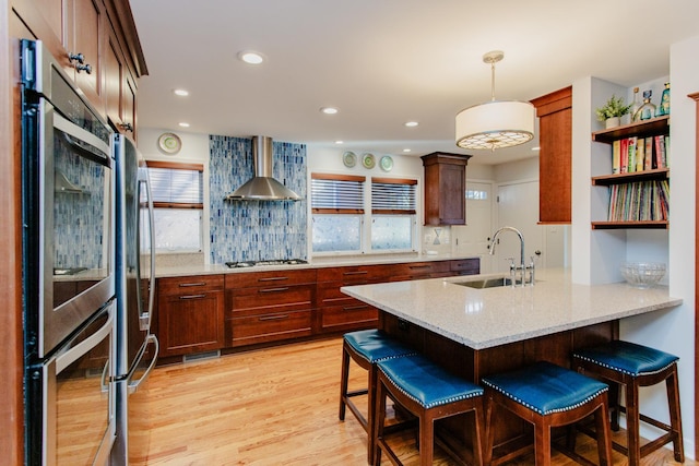 kitchen featuring sink, stainless steel appliances, decorative light fixtures, wall chimney exhaust hood, and light hardwood / wood-style floors