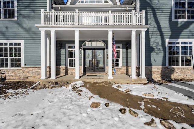 snow covered property entrance with a balcony, stone siding, and french doors