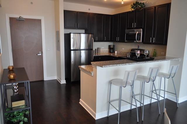 kitchen with appliances with stainless steel finishes, a kitchen breakfast bar, kitchen peninsula, light stone countertops, and dark wood-type flooring