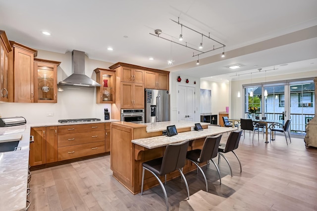 kitchen featuring pendant lighting, a center island, wall chimney exhaust hood, light wood-type flooring, and appliances with stainless steel finishes