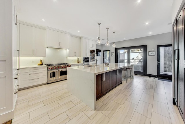 kitchen featuring range with two ovens, pendant lighting, light countertops, white cabinets, and an island with sink