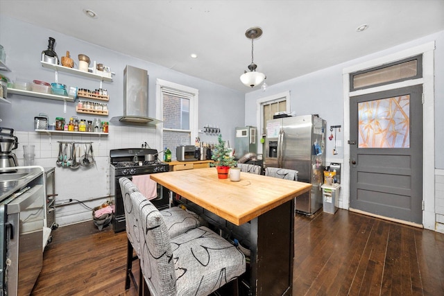 kitchen featuring stainless steel fridge, dark hardwood / wood-style flooring, black range with gas stovetop, wall chimney range hood, and decorative light fixtures