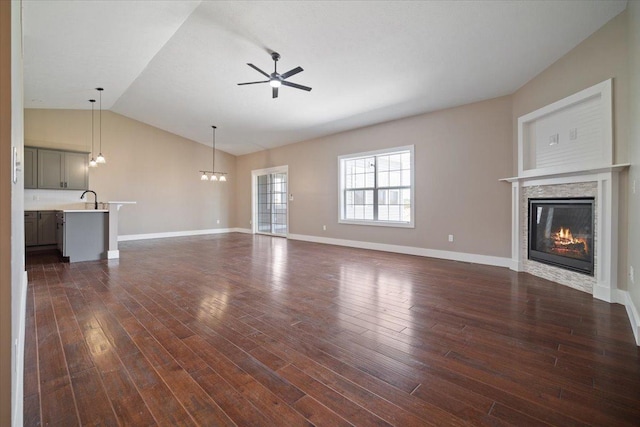 unfurnished living room featuring ceiling fan with notable chandelier, dark wood finished floors, baseboards, vaulted ceiling, and a glass covered fireplace