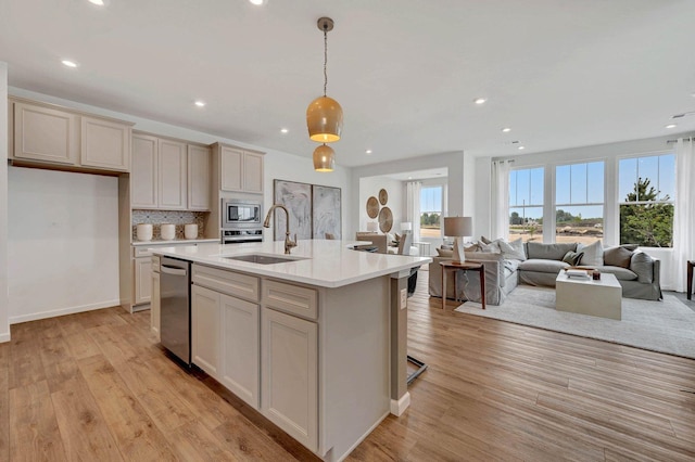 kitchen featuring appliances with stainless steel finishes, a kitchen island with sink, sink, light hardwood / wood-style flooring, and hanging light fixtures