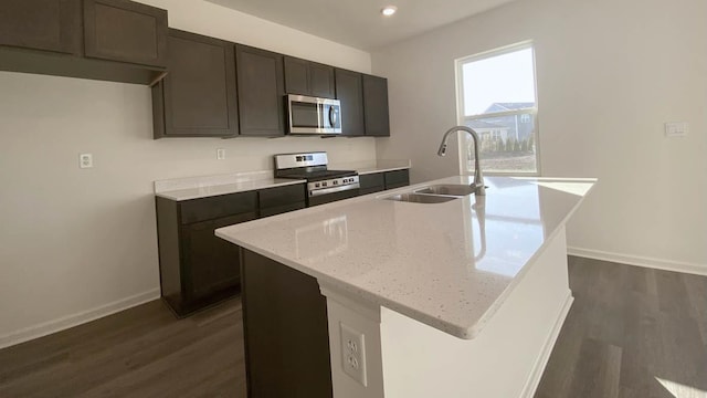 kitchen with a center island with sink, sink, light stone countertops, appliances with stainless steel finishes, and dark hardwood / wood-style flooring