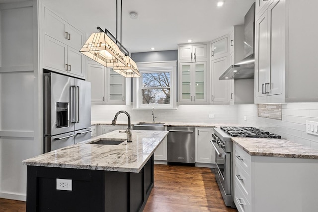 kitchen featuring premium appliances, sink, wall chimney range hood, decorative light fixtures, and a center island with sink