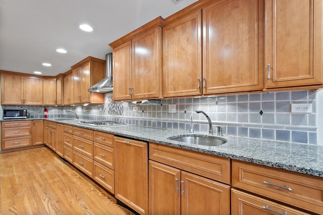 kitchen featuring light stone counters, light wood-style flooring, appliances with stainless steel finishes, a sink, and wall chimney range hood