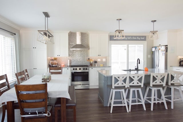 kitchen with wall chimney exhaust hood, stainless steel appliances, a kitchen island with sink, pendant lighting, and white cabinets