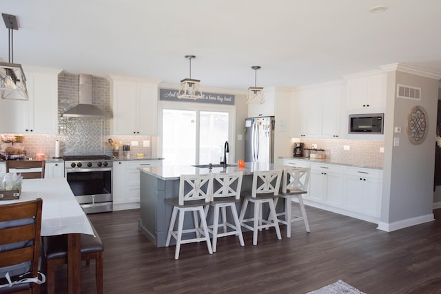 kitchen featuring pendant lighting, white cabinets, wall chimney exhaust hood, an island with sink, and stainless steel appliances