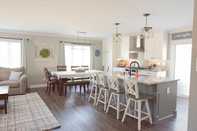 kitchen featuring a kitchen bar, light stone countertops, wall chimney range hood, pendant lighting, and white cabinetry