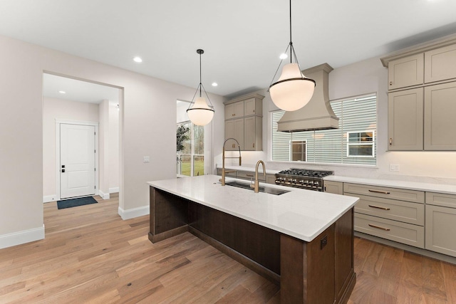 kitchen with light hardwood / wood-style floors, custom exhaust hood, pendant lighting, stainless steel stove, and an island with sink