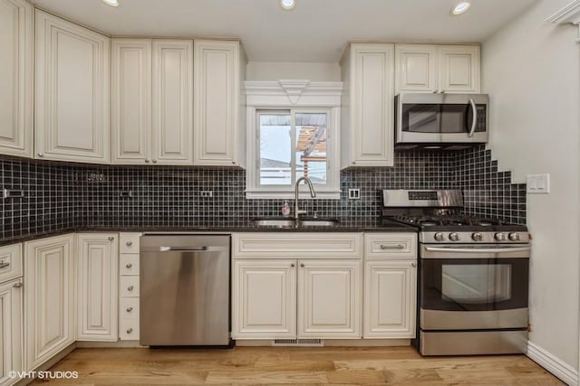 kitchen with appliances with stainless steel finishes, tasteful backsplash, sink, dark stone countertops, and light wood-type flooring