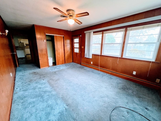 unfurnished bedroom with baseboard heating, ceiling fan, wooden walls, and light colored carpet