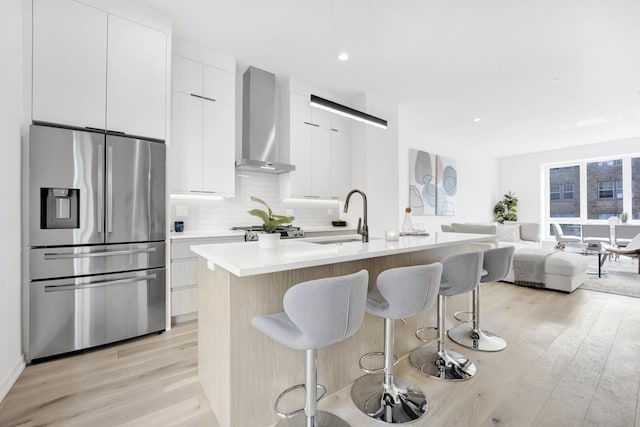 kitchen with a kitchen island with sink, white cabinets, wall chimney range hood, sink, and stainless steel refrigerator with ice dispenser