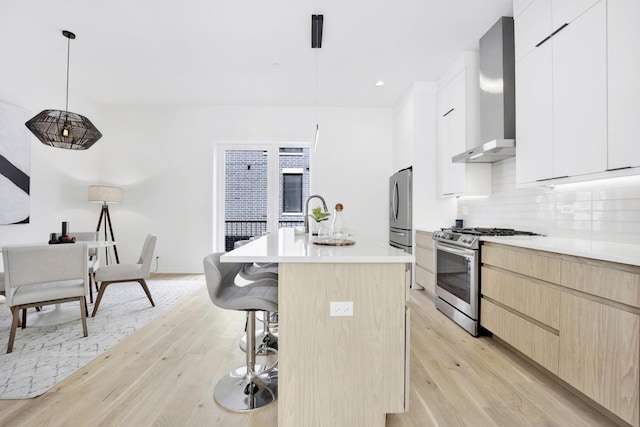 kitchen with a kitchen island with sink, hanging light fixtures, wall chimney exhaust hood, white cabinetry, and stainless steel appliances