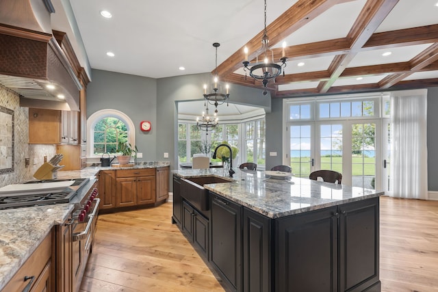 kitchen featuring pendant lighting, beamed ceiling, an island with sink, sink, and light stone counters