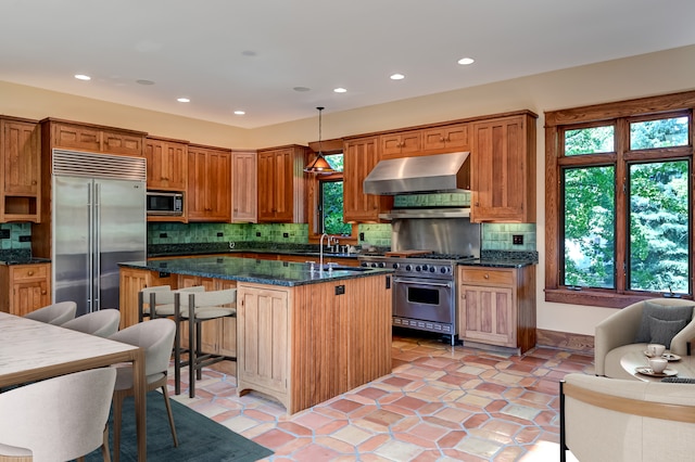 kitchen with high end appliances, tasteful backsplash, hanging light fixtures, a kitchen island with sink, and wall chimney range hood