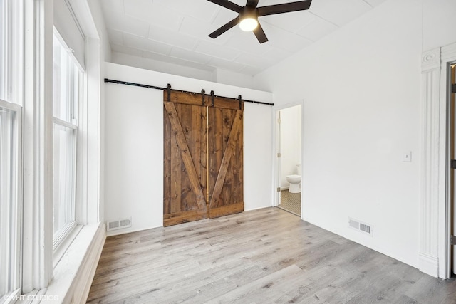 unfurnished bedroom featuring light wood-type flooring, a barn door, ensuite bath, and ceiling fan