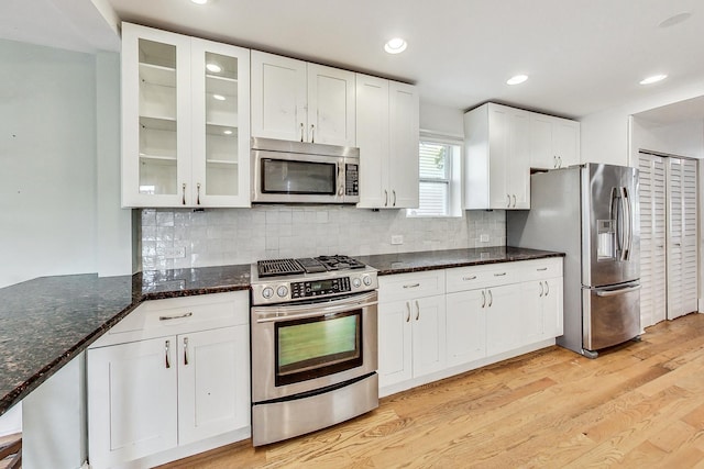 kitchen featuring white cabinets, light wood-style flooring, glass insert cabinets, appliances with stainless steel finishes, and dark stone countertops