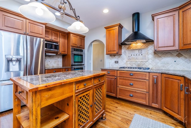 kitchen with wall chimney exhaust hood, hanging light fixtures, tasteful backsplash, appliances with stainless steel finishes, and light wood-type flooring