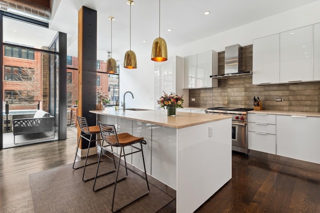 kitchen with wall chimney range hood, pendant lighting, a kitchen island with sink, designer range, and white cabinets