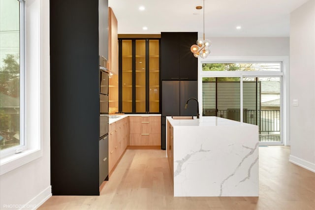 kitchen with decorative light fixtures, light hardwood / wood-style flooring, light brown cabinets, light stone countertops, and a kitchen island with sink