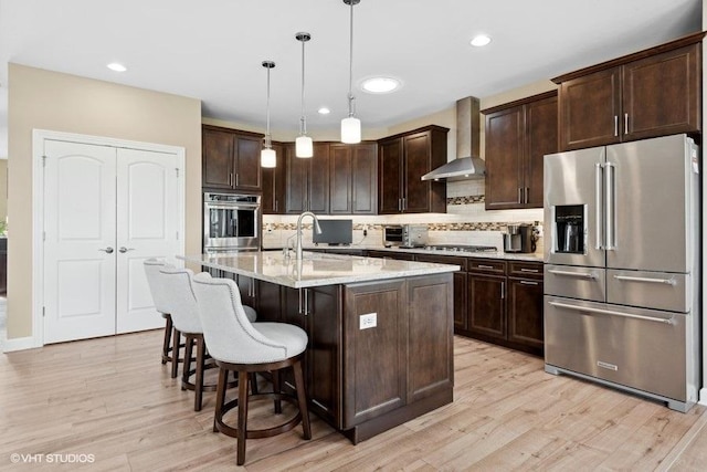 kitchen with a center island with sink, wall chimney range hood, sink, hanging light fixtures, and stainless steel appliances