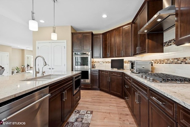 kitchen with wall chimney range hood, hanging light fixtures, sink, appliances with stainless steel finishes, and tasteful backsplash