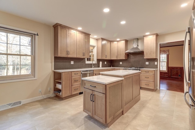 kitchen with light brown cabinetry, stainless steel appliances, sink, wall chimney range hood, and a kitchen island