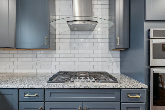 kitchen with light stone countertops, wall chimney range hood, backsplash, and appliances with stainless steel finishes