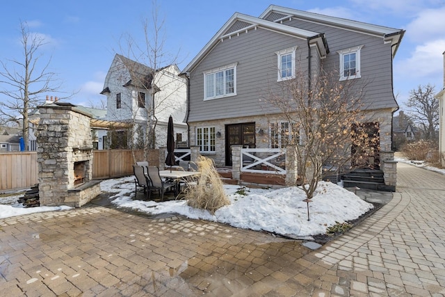 snow covered back of property with stone siding, fence, an outdoor stone fireplace, and a patio