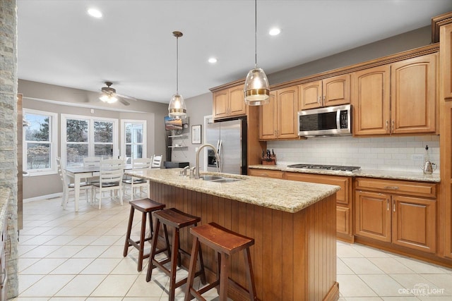 kitchen with light stone countertops, pendant lighting, a center island with sink, appliances with stainless steel finishes, and ceiling fan