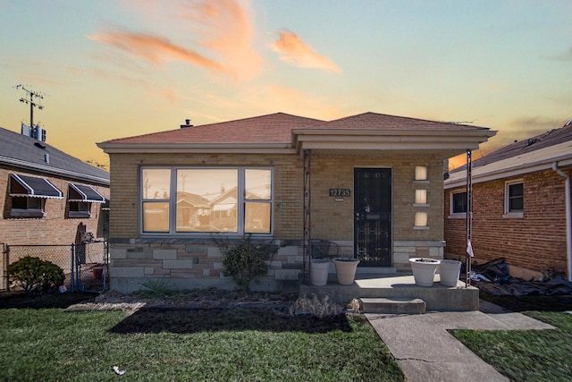 bungalow with stone siding, a lawn, fence, and roof with shingles