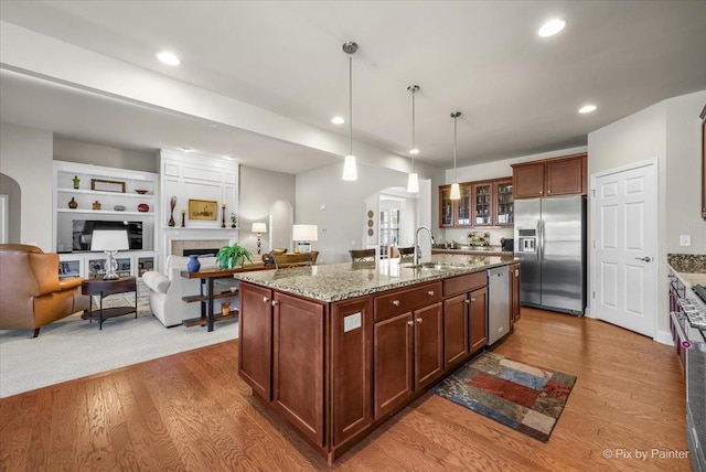 kitchen with appliances with stainless steel finishes, built in shelves, a kitchen island with sink, pendant lighting, and a tiled fireplace