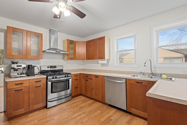 kitchen with stainless steel appliances, ceiling fan, sink, wall chimney range hood, and light hardwood / wood-style flooring