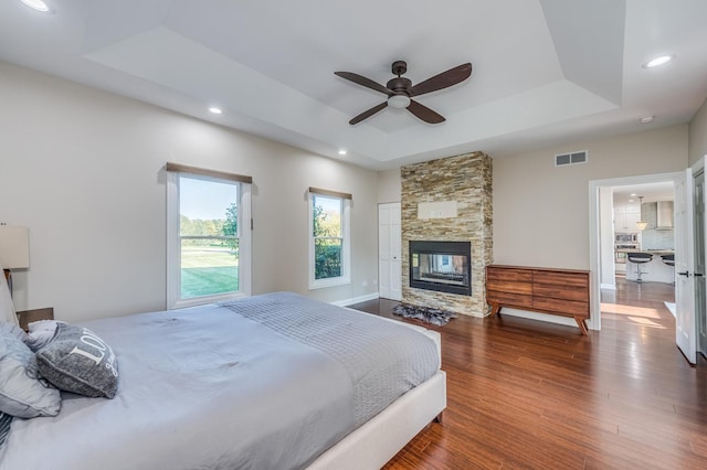 bedroom featuring dark hardwood / wood-style flooring, a tray ceiling, a stone fireplace, and ceiling fan