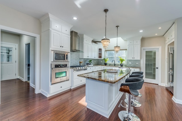 kitchen with white cabinets, wall chimney range hood, appliances with stainless steel finishes, stone countertops, and a kitchen island