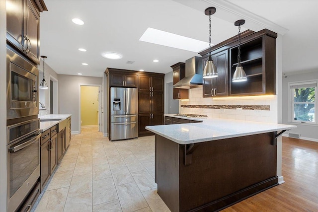 kitchen featuring a skylight, wall chimney exhaust hood, a kitchen breakfast bar, decorative light fixtures, and appliances with stainless steel finishes