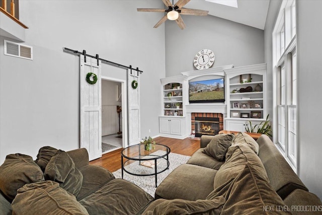 living room featuring ceiling fan, a brick fireplace, a barn door, high vaulted ceiling, and hardwood / wood-style flooring