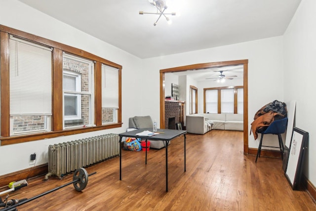 home office featuring hardwood / wood-style floors, ceiling fan with notable chandelier, a brick fireplace, and radiator