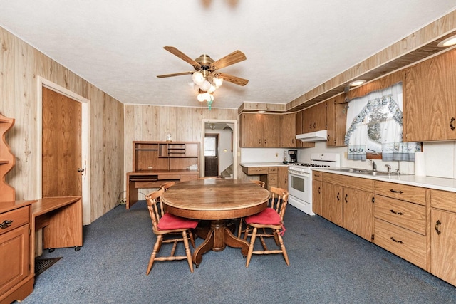 kitchen with sink, white range with gas stovetop, dark colored carpet, wooden walls, and ceiling fan