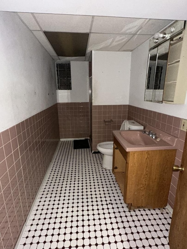 bathroom with a drop ceiling, vanity, tile walls, and toilet