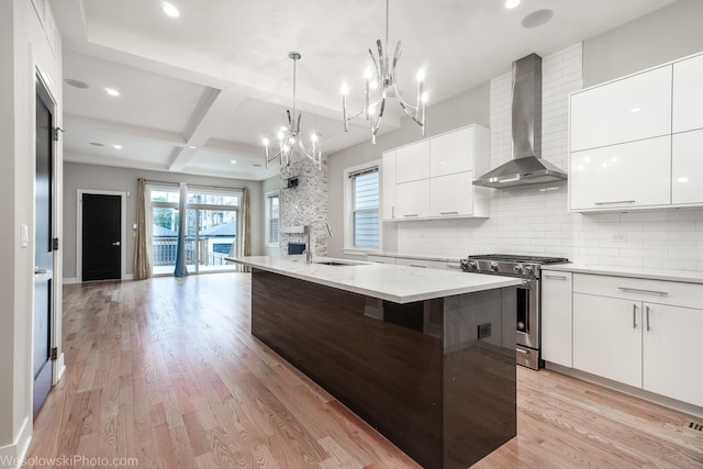 kitchen with coffered ceiling, wall chimney exhaust hood, stainless steel gas stove, an island with sink, and beamed ceiling