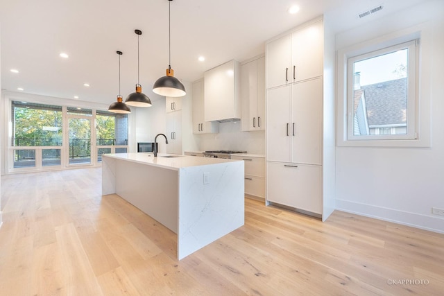 kitchen featuring custom range hood, sink, a center island with sink, white cabinetry, and hanging light fixtures