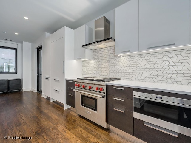 kitchen featuring wall chimney exhaust hood, dark wood-type flooring, white cabinetry, decorative backsplash, and premium range