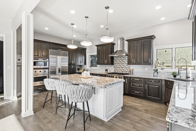 kitchen featuring sink, light stone counters, a kitchen island, stainless steel appliances, and wall chimney range hood