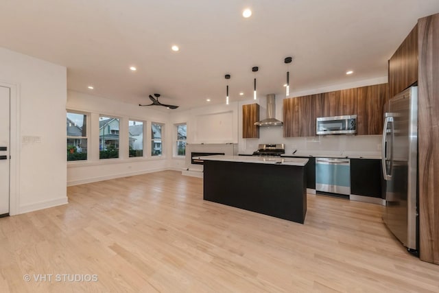 kitchen with decorative light fixtures, stainless steel appliances, wall chimney range hood, a kitchen island, and ceiling fan