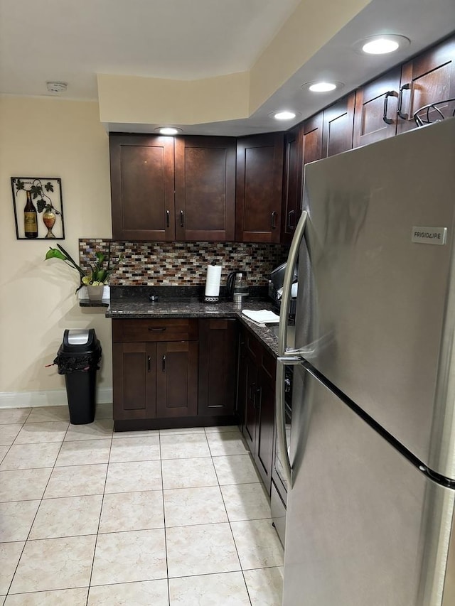 kitchen with dark brown cabinetry, backsplash, dark stone countertops, stainless steel fridge, and light tile patterned floors