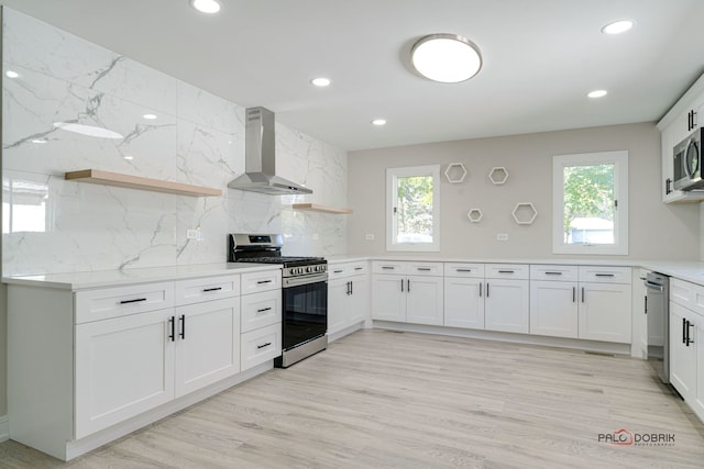 kitchen featuring appliances with stainless steel finishes, tasteful backsplash, wall chimney range hood, light hardwood / wood-style floors, and white cabinetry